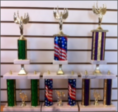 Three trophies varing in size. Left= white base with green columns and vicotry topper and trim topper on bottom. Middle = Victory topper on U.S.A. flag post with white bases and trim topper on bottom, right = victory topper on blue/gold columns with white